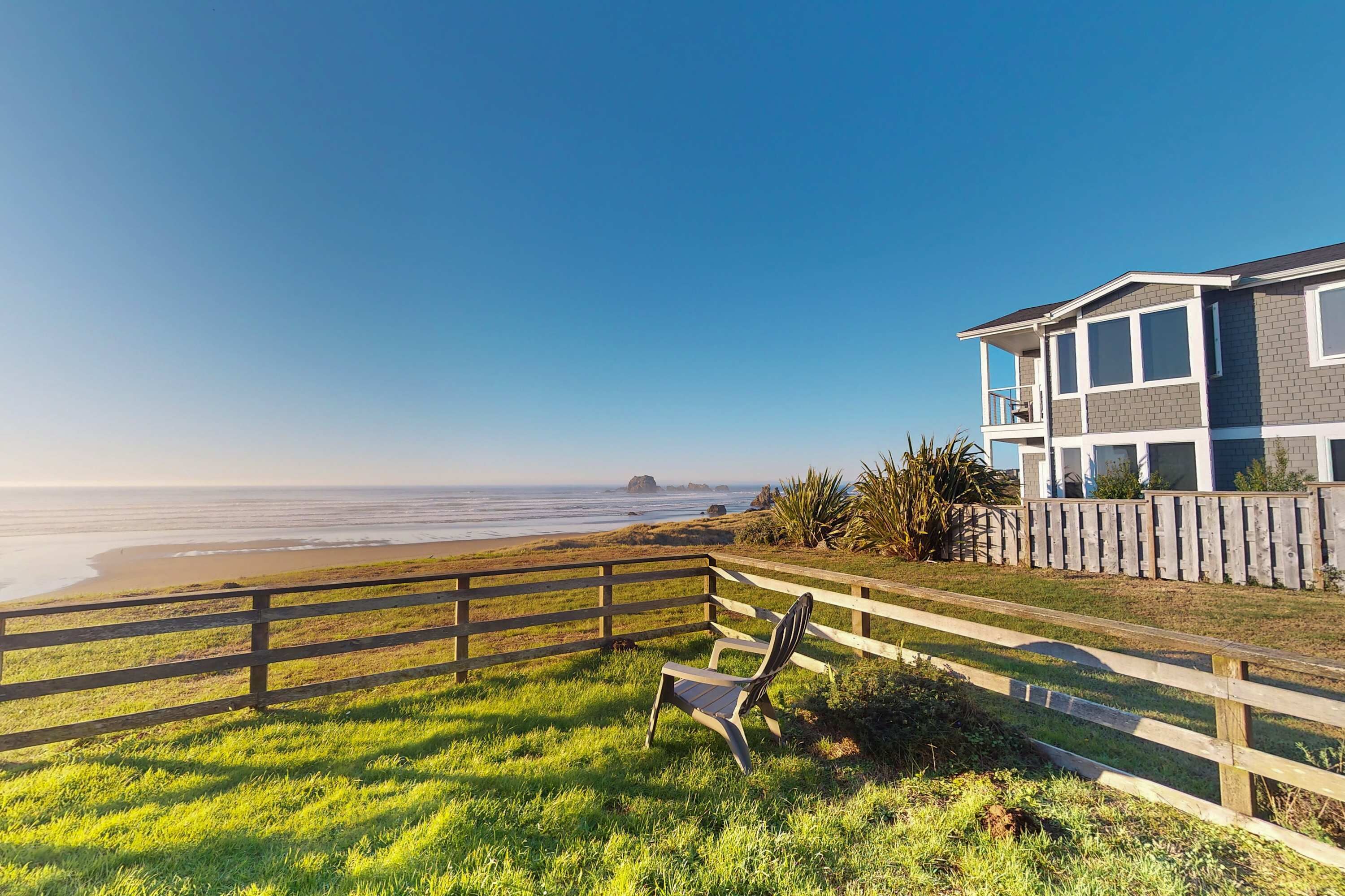Une vue sur la plage avec une clôture en bois, une chaise et une maison dotée d’un balcon.