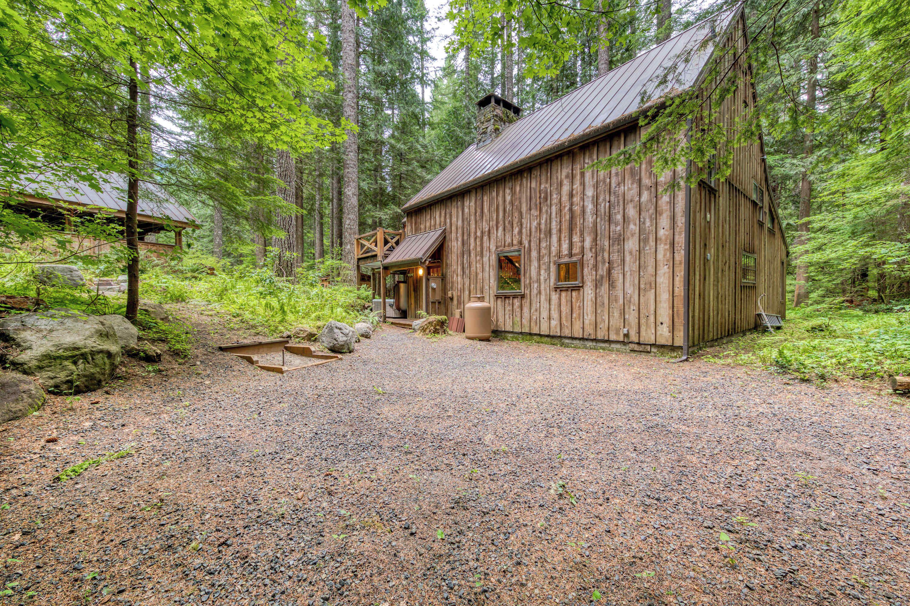 Un chalet rustique en bois, avec une allée en gravier, entouré d’une forêt dense.