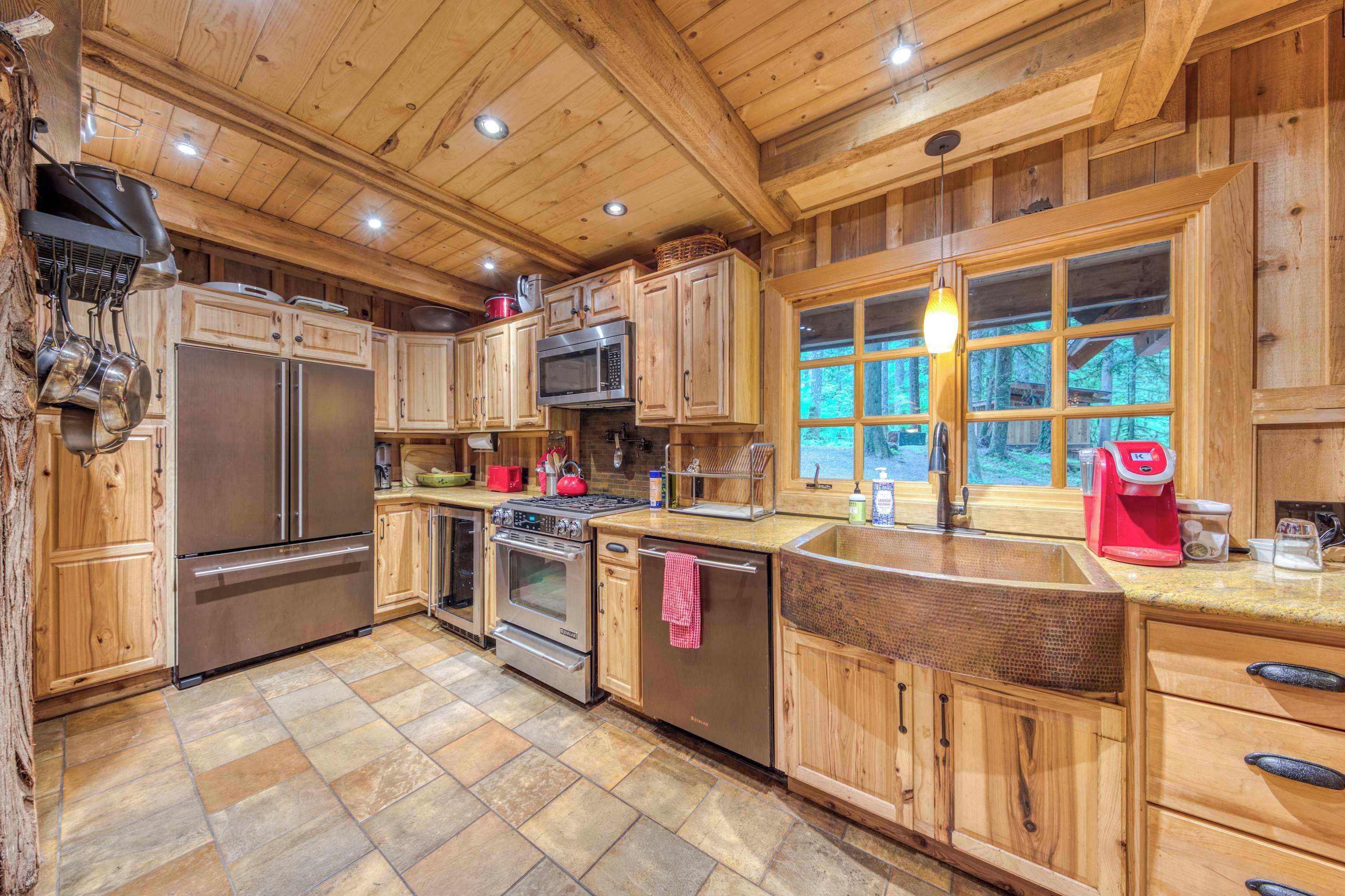 Une cuisine rustique, située dans une cabane en bois, avec des appareils en acier inoxydable, une grande fenêtre et un sol carrelé en pierre.