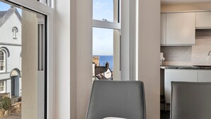 A dining area with a table set for two, a view of the sea through large windows, and a modern kitchen in the background. - Sea Steam Apartment - Close to Village and Beach (Saundersfoot)