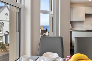 Ein Essbereich mit Tisch für zwei, Meerblick durch große Fenster und eine moderne Küche im Hintergrund.