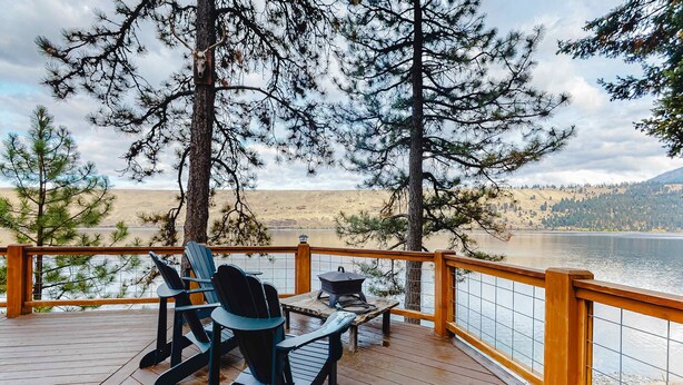 A deck with chairs and a table, overlooking a lake and forested hills.