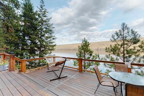 A wooden deck with a hammock, chairs, and a table overlooking a lake with trees and hills.