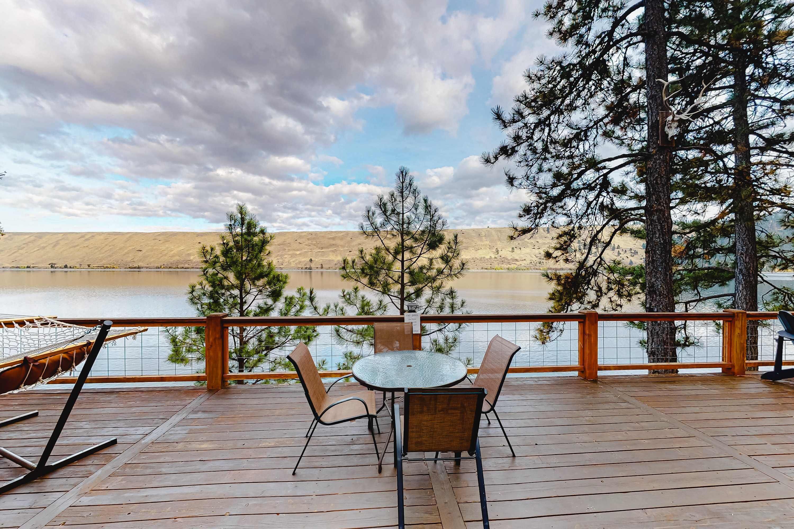 A wooden deck with a table and chairs, overlooking a body of water and a hilly landscape.