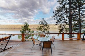 A wooden deck with a table and chairs, overlooking a body of water and a hilly landscape.
