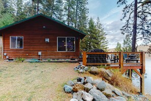 A wooden cabin with a deck, a grassy yard, and a rocky shoreline.