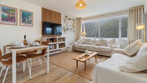 Ein modernes Wohnzimmer mit Essbereich, weißen Möbeln, einem Flachbildfernseher und großen Fenstern.