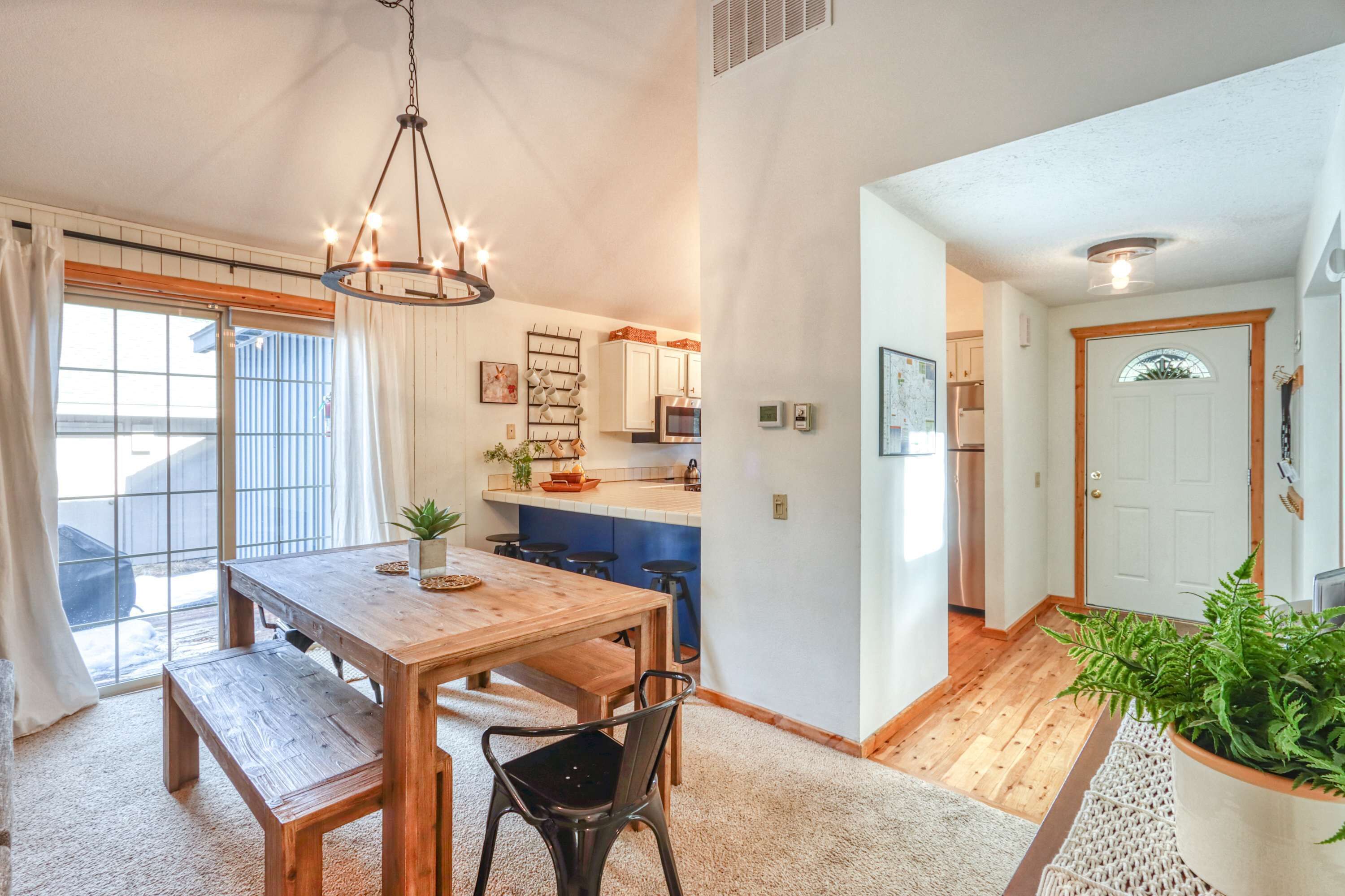 A dining area with a wooden table and chairs, a hanging light fixture, and a view of the kitchen.