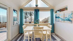 A dining area with a table and chairs, large windows with curtains, and a view of trees.