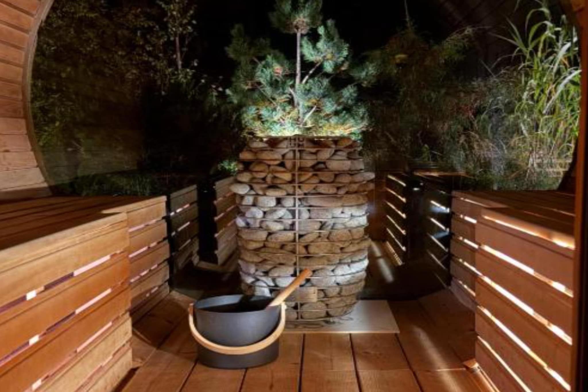 An indoor sauna with a stone wall, wooden benches, and a wooden bucket.