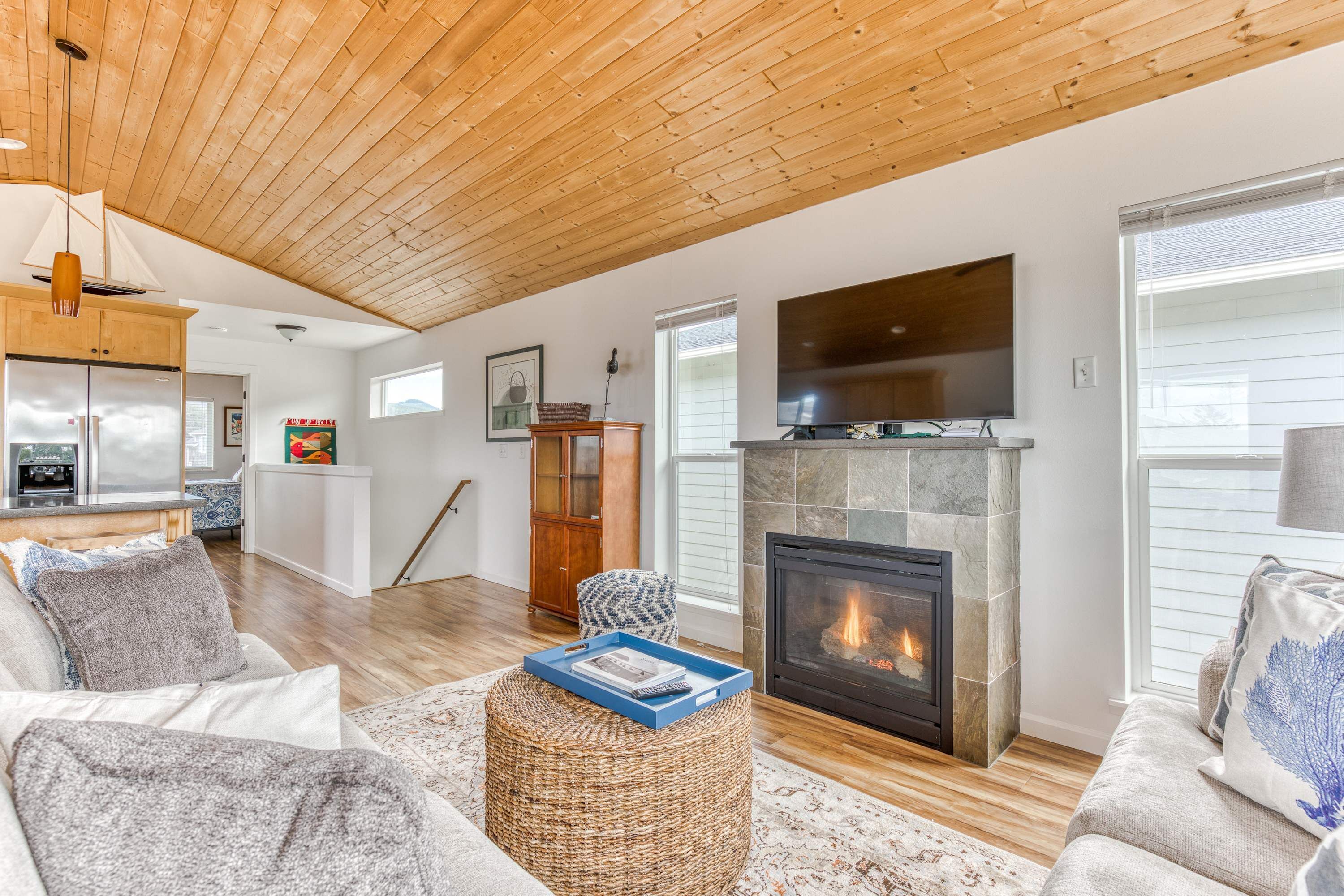 A living room with a fireplace, wooden ceiling, and a dining area with a kitchen in the background.