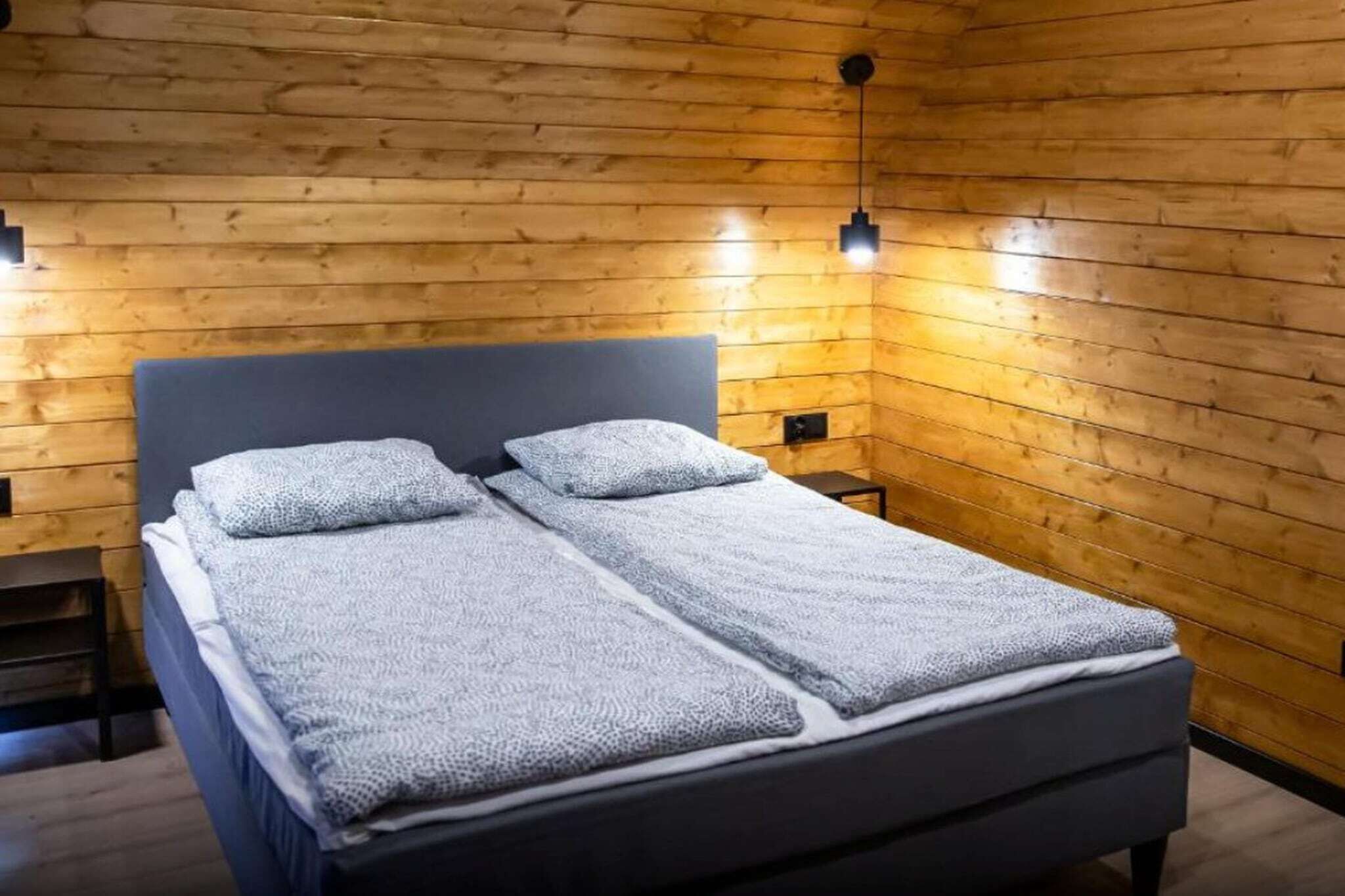 Un lit double avec une tête de lit grise et deux oreillers, placé contre un mur en bois avec un luminaire suspendu.