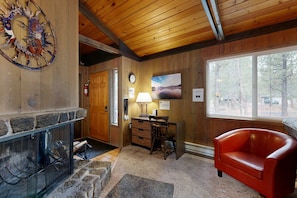 A cozy room with a stone fireplace, wooden walls, a red leather chair, and a large window. - Comfortable Family-Friendly Sunriver Retreat with Deck & Grill - Free SHARC Access (Sunriver)