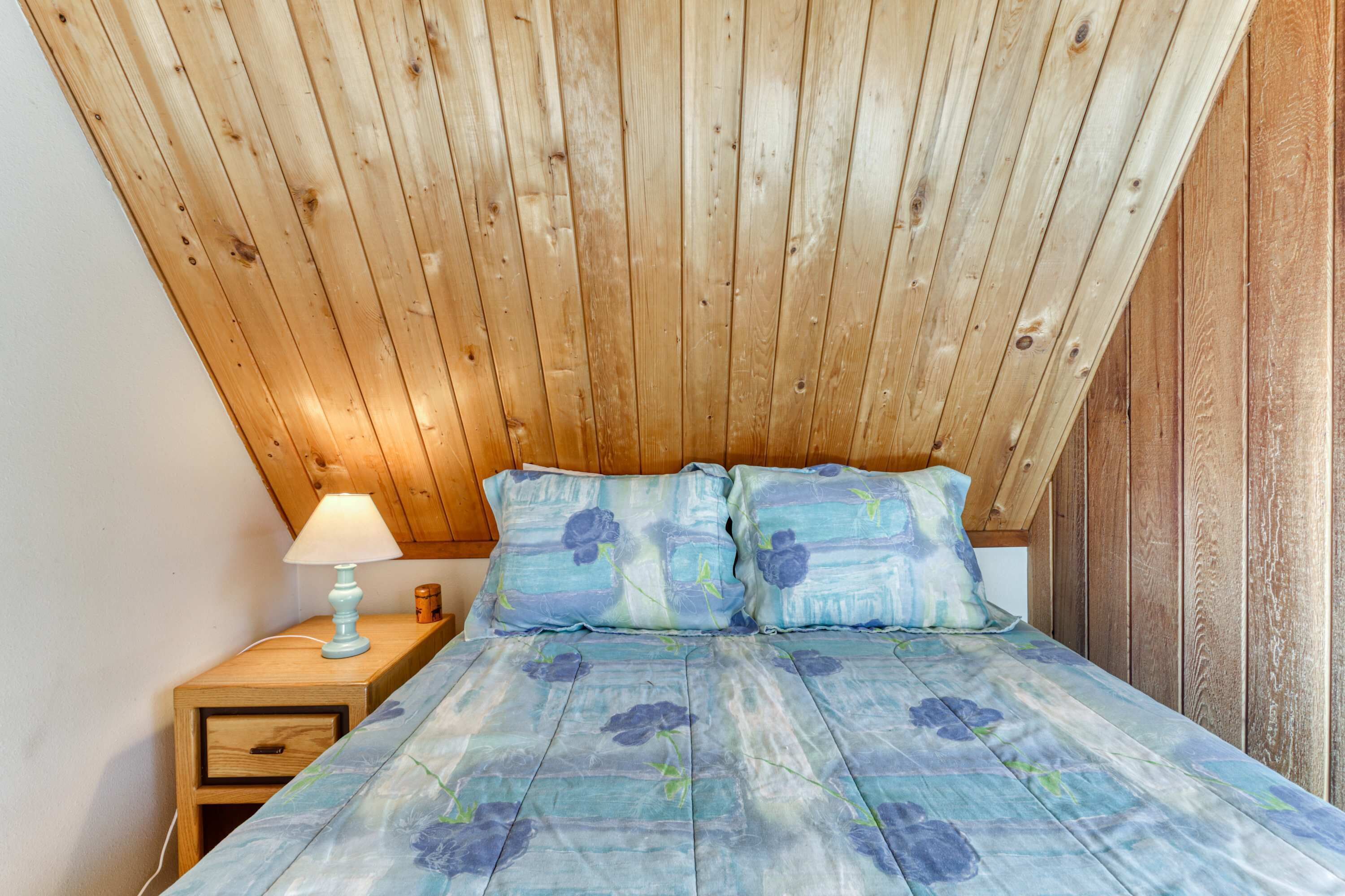 A bedroom with a wooden ceiling, a bed with floral bedding, a bedside table with a lamp, and a nightstand with a drawer.