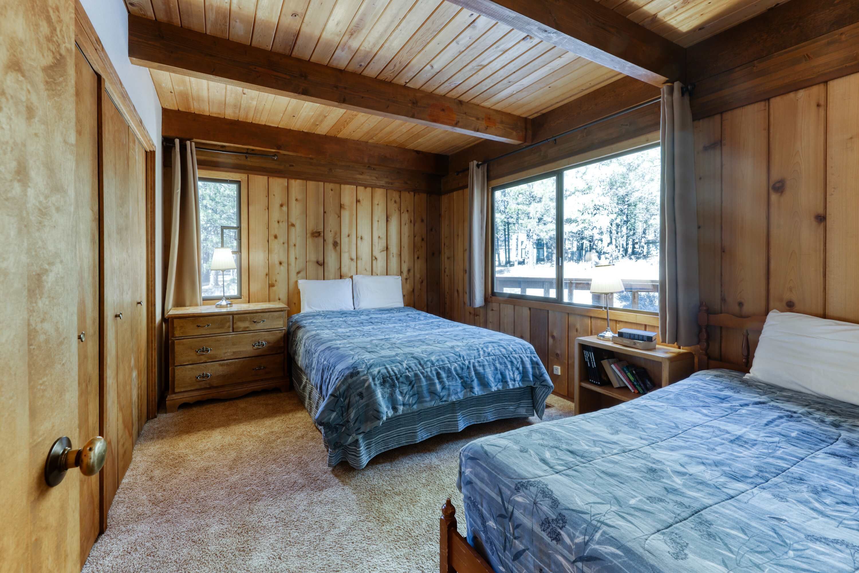 Een slaapkamer in een houten chalet met twee bedden, een nachtkastje, een dressoir en een raam met uitzicht op bomen.