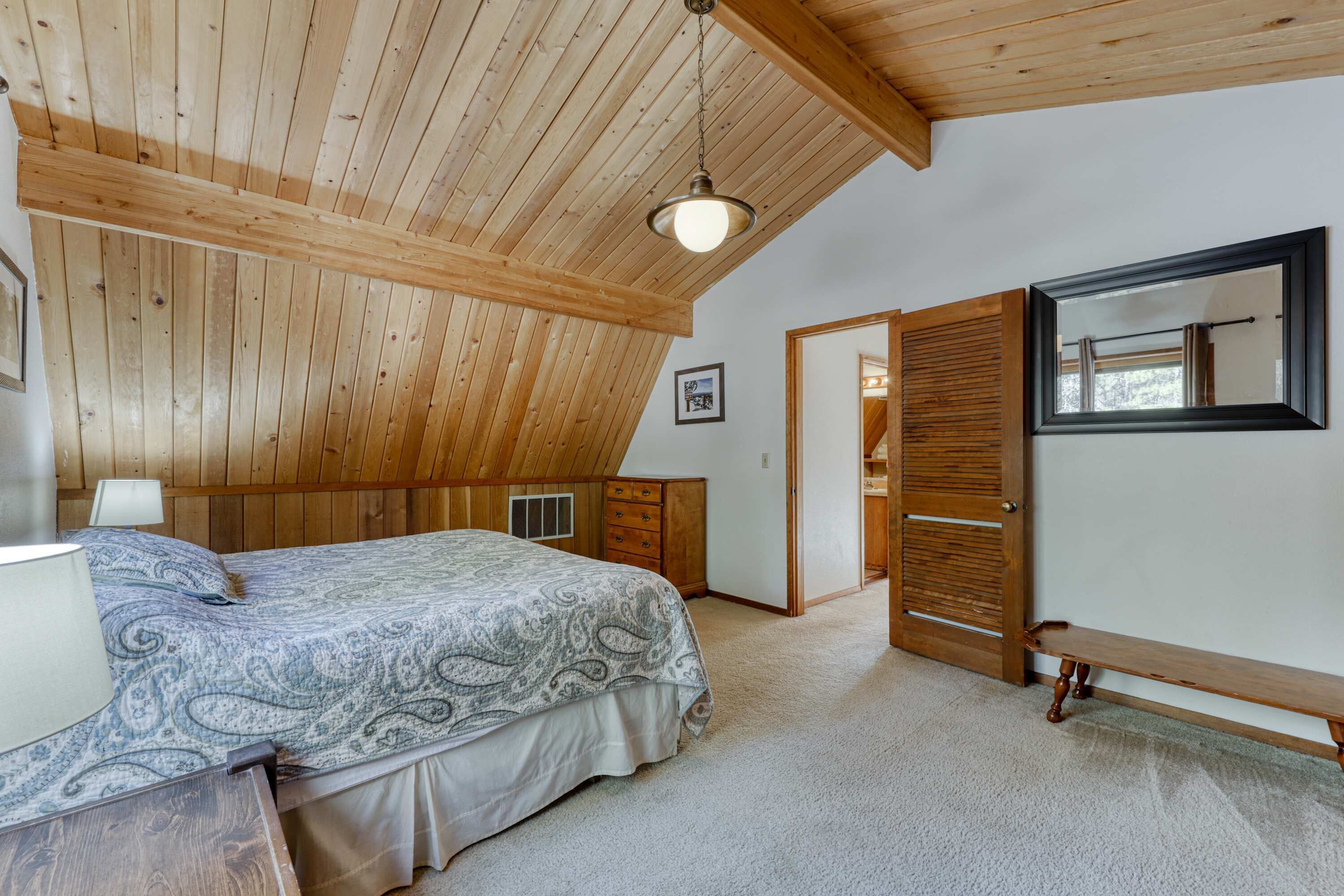 Une chambre avec un plafond en pente, un lit recouvert d’une couvre-lit à motifs, une lampe de chevet, un banc en bois et un miroir accroché au mur.