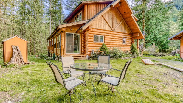 A log cabin with a glass dining table and chairs, surrounded by a grassy yard and dense forest. - Woodland home near golf & ski slopes with wraparound deck - dog-friendly (Welches)