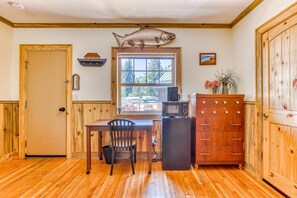 A kitchen with wooden floors, a wooden desk, a chair, a refrigerator, a microwave, a fish mounted above the window, and a wooden cabinet.