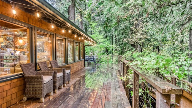 A wooden deck with wicker furniture and a glass enclosure, surrounded by dense forest. - Charming Home with Free WiFi, washer&dryer, Wood Fireplace, and Furnished Deck (Sandy)