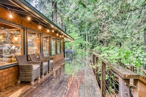 A wooden deck with wicker furniture and a glass enclosure, surrounded by dense forest. - Charming Home with Free WiFi, washer&dryer, Wood Fireplace, and Furnished Deck (Sandy)