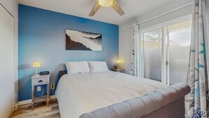A bedroom with a bed, a nightstand, a ceiling fan, a window with curtains, and a wall painting. - Family Friendly Coastal Getaway Townhome Near Beach & Downtown (Long Beach)