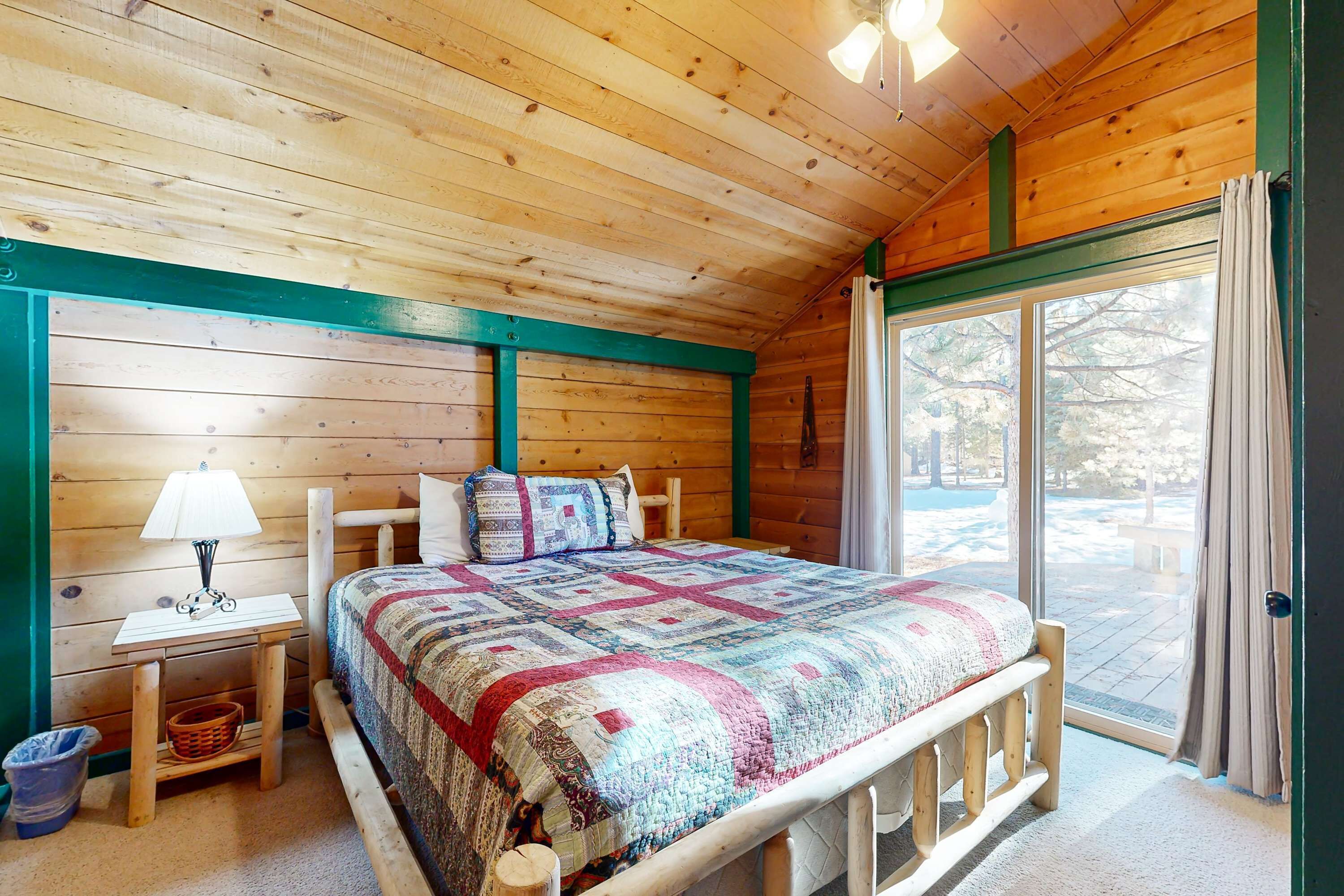 Een houten slaapkamer met een bed, nachtkastje, lamp, raam en uitzicht op de natuur.