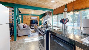 A modern kitchen with a built-in oven and dishwasher, a stone fireplace, and a dining area with a sofa and coffee table. - Cabin with Eight SHARC Passes, Access to Shared Pools, Hot Tub & Other Amenities (Sunriver)