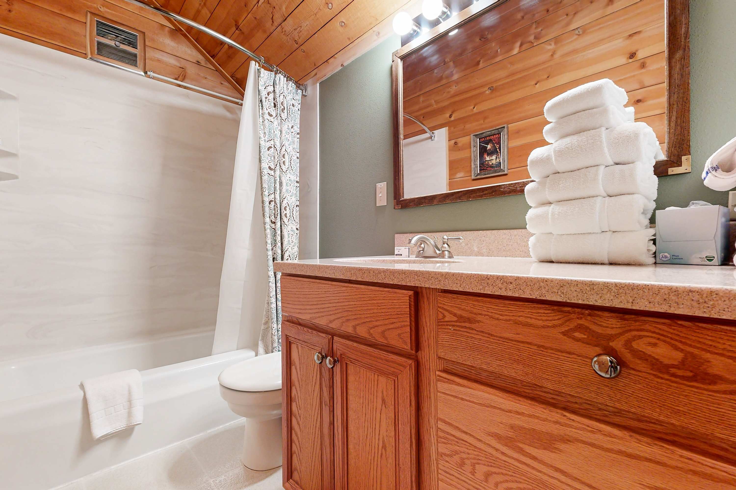A bathroom with a wooden vanity, a stack of towels, a shower curtain, and a mirror.