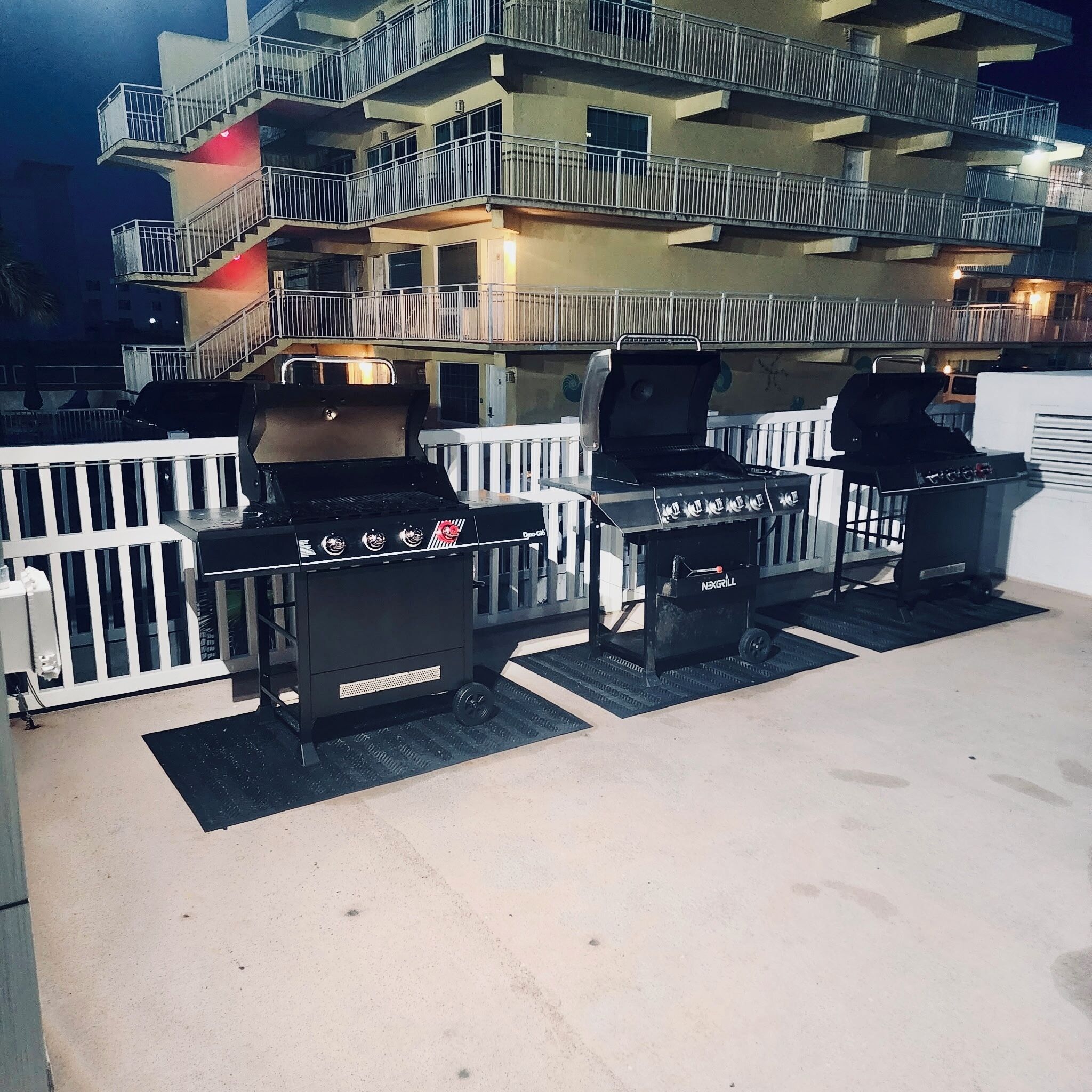 An outdoor barbecue area with two grills, a white fence, and a multi-story building in the background.