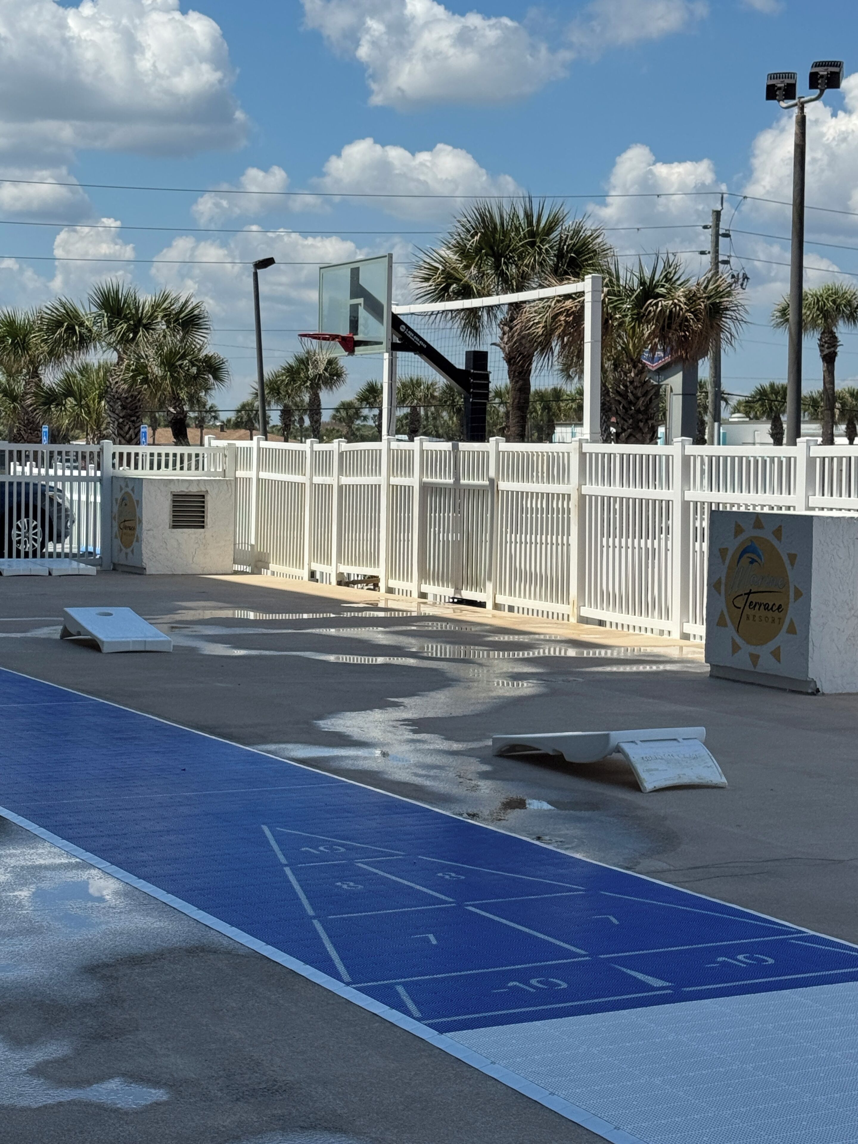 Ein Sportplatz mit einem Basketballkorb, einem weißen Zaun und im Hintergrund Palmen.