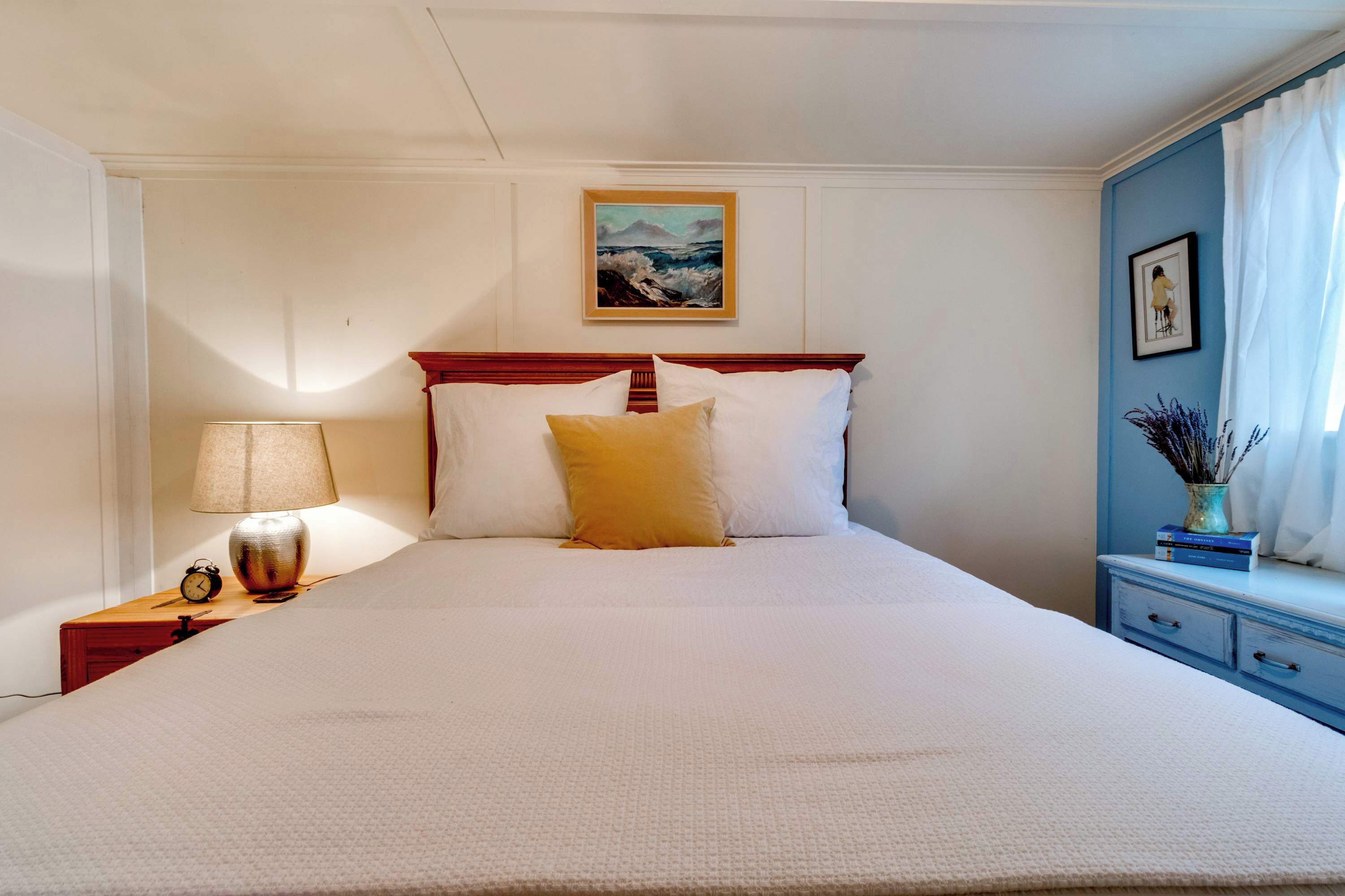 Una cama bien hecha con almohadas blancas y amarillas, una lámpara de noche y un cuadro en la pared.