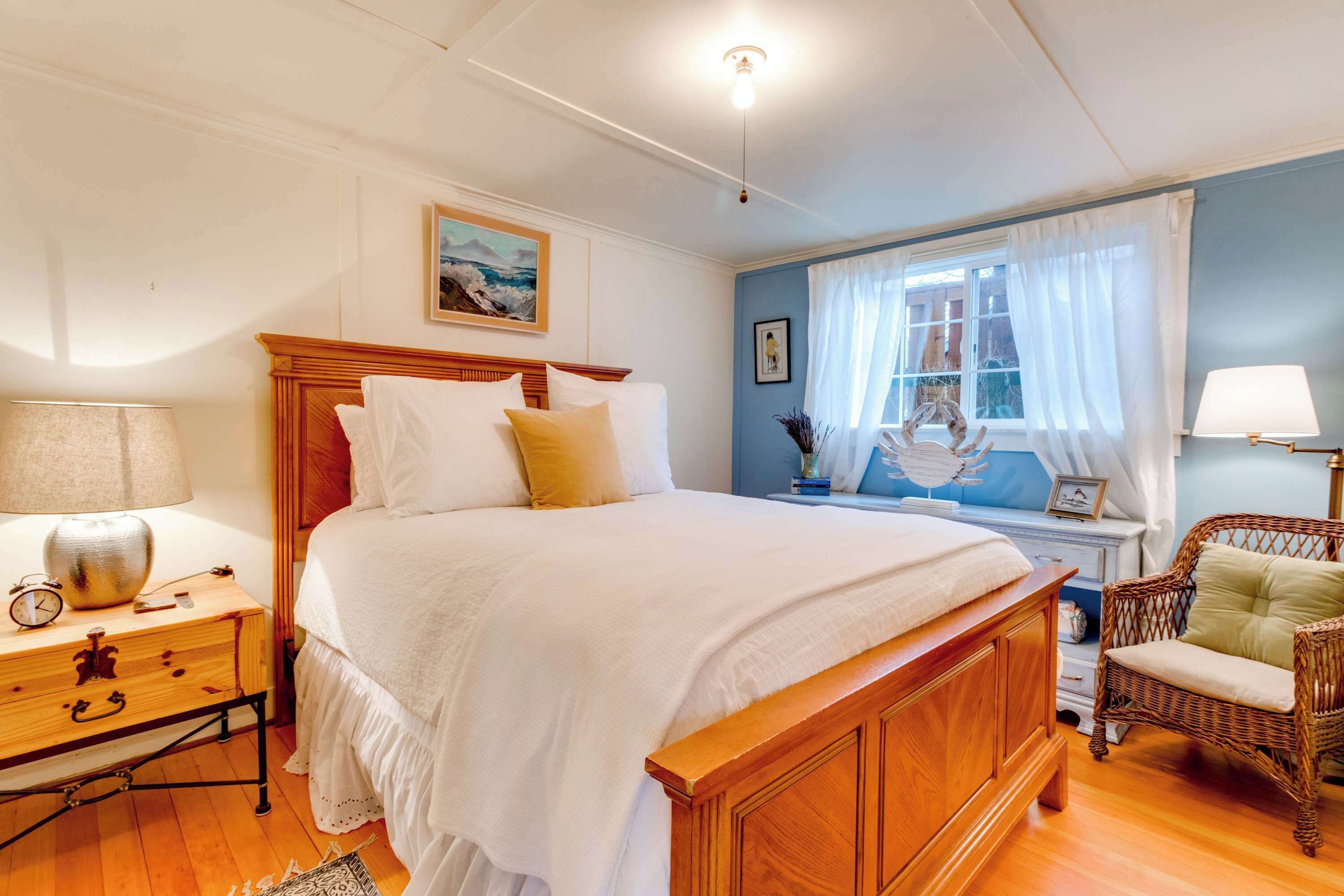A bedroom with a wooden bed, a nightstand with a lamp, a wicker chair, and a window with curtains.