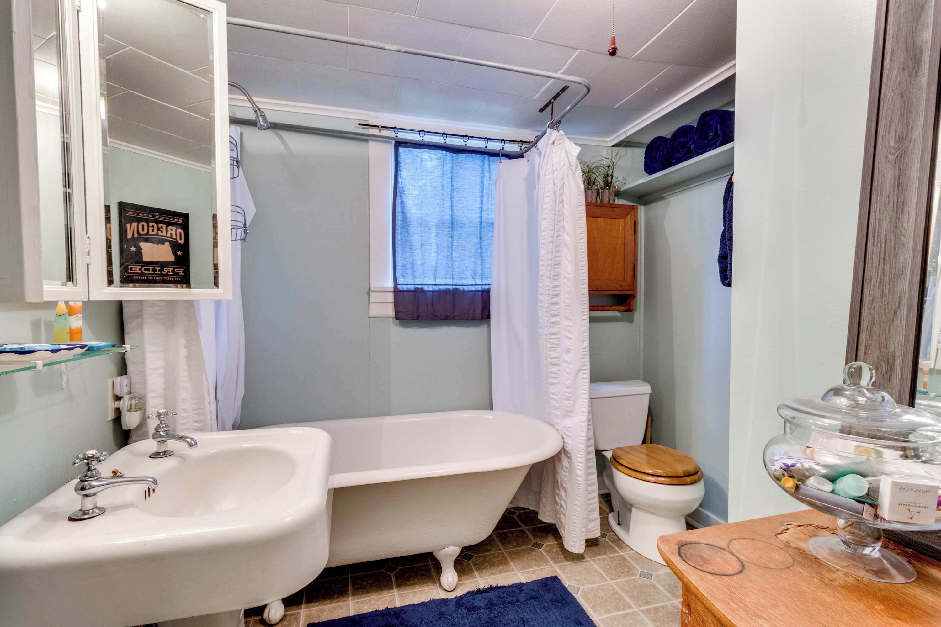 A bathroom with a freestanding bathtub, a white sink, a wooden toilet, and a glass jar of toiletries on a wooden table.