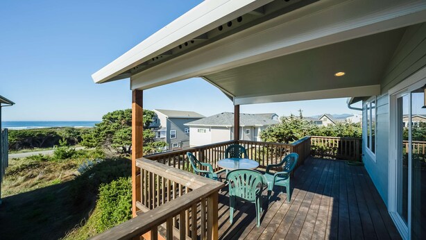 Eine Holzterrasse mit überdachter Sitzgruppe, mit Blick auf einen Strand und Wohnhäuser.