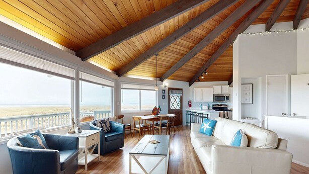 Amplia sala de estar con techo de madera, ventanas grandes, sofá blanco, sillón azul y una mesa de comedor.