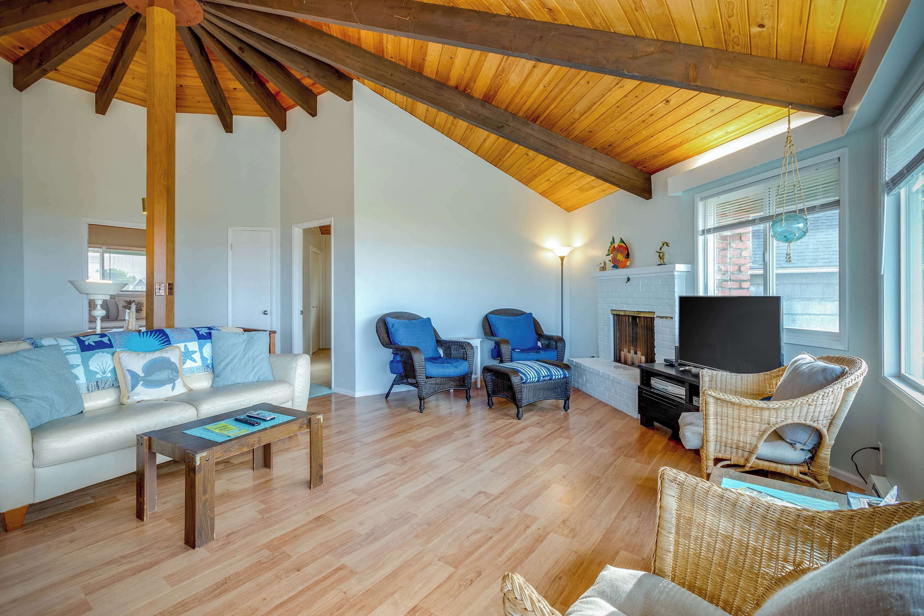 A spacious living room with a wooden ceiling, a white sofa with blue cushions, wicker chairs, a television, and a fireplace.