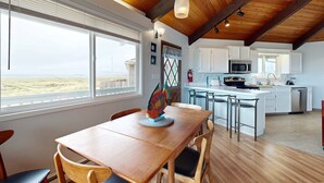 Un comedor con mesa y sillas de madera, una cocina con armarios blancos, y vista a la playa.