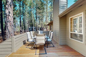 A wooden deck with a table and chairs, surrounded by tall trees. - Cozy home with private hot tub, patio dining, gas fireplace and full kitchen (Sunriver)