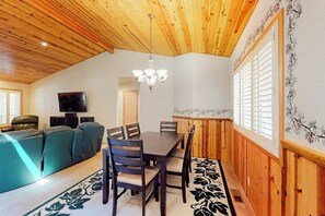 A cozy living area with a wooden ceiling, a dining table with chairs, a sofa, and a TV mounted on the wall. - Cozy home with private hot tub, patio dining, gas fireplace and full kitchen (Sunriver)