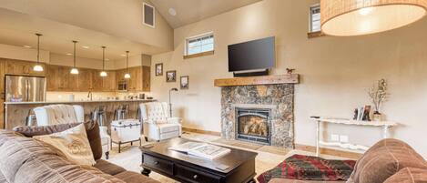 A spacious living room with a fireplace, a television mounted above it, and a kitchen area in the background.