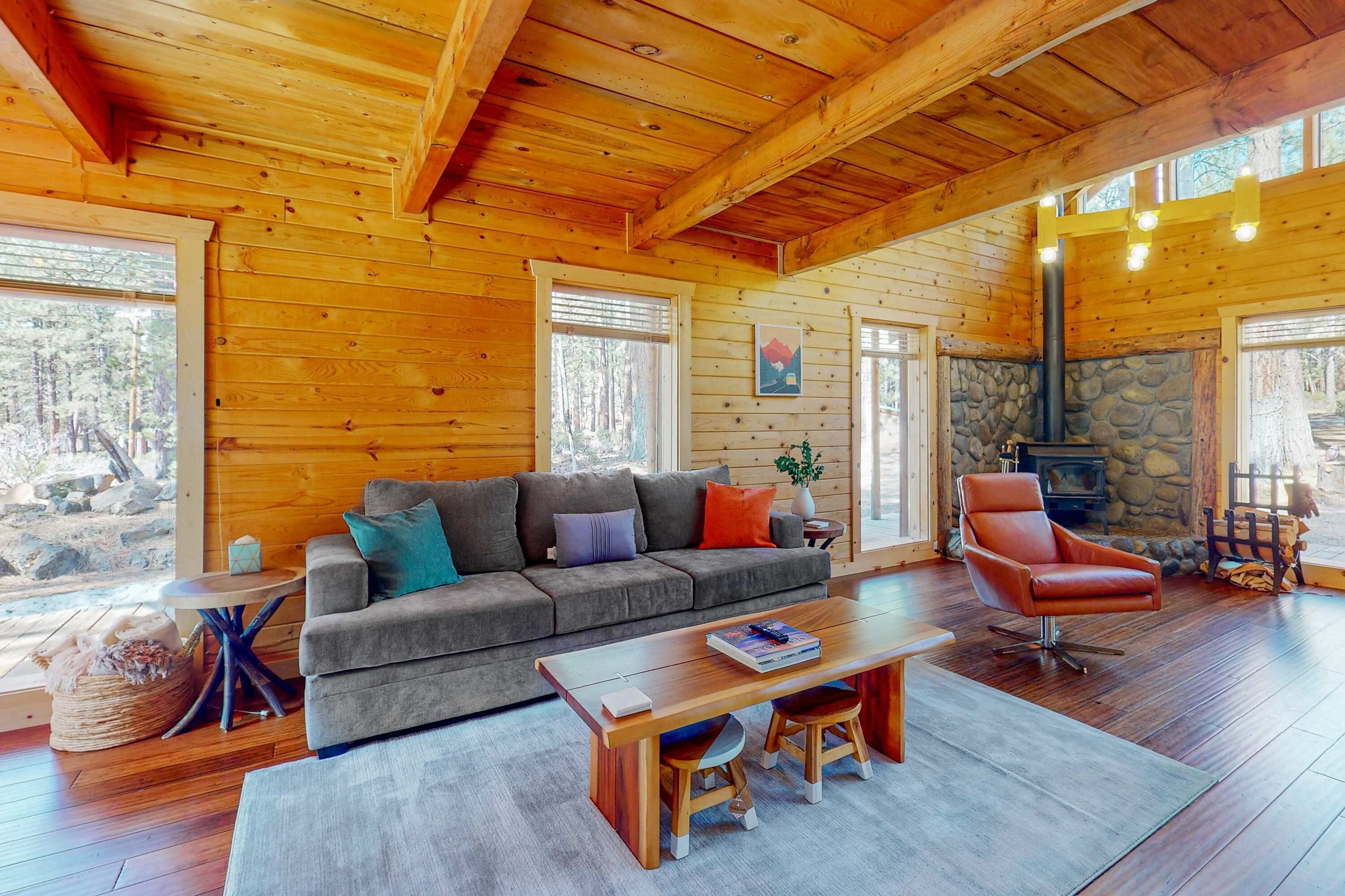 Ein gemütliches Blockhaus-Interieur mit Holzbalkendecke, Kamin aus Stein, einer Couch, einem Ledersessel und einem Esstisch.