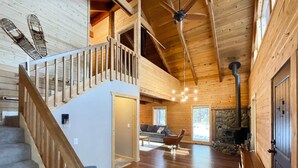Ein Holzblockhaus-Innenraum mit Treppe, einem Wohnbereich mit Sofa und Kamin sowie einem Deckenventilator.