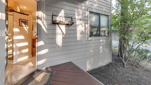 Une entrée de maison avec une porte en bois, un paillasson, un luminaire fixé au mur et un petit arbre.