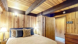 A bedroom with a wooden ceiling, a bed with patterned bedding, a nightstand with a lamp, and a wardrobe. - Charming 2-story chalet with deck, wood stove, Deschutes River access (Sunriver)