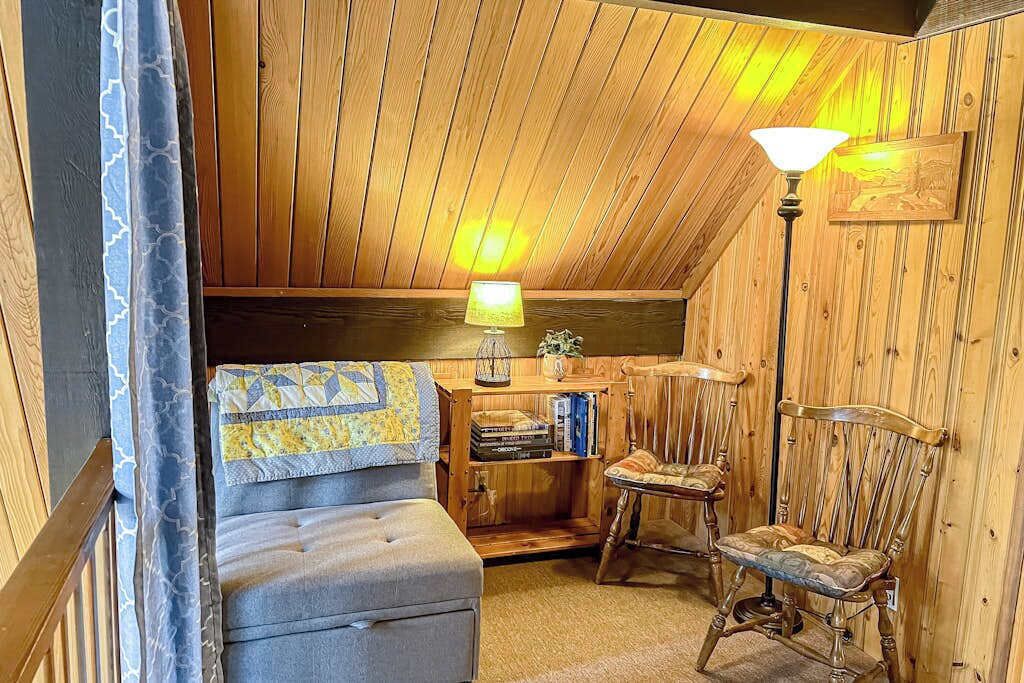 Una habitación acogedora con techo de madera, un banco acolchado, una silla de madera, una lámpara y una estantería.
