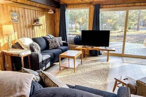 Ein gemütliches Wohnzimmer mit einer Couch, einem Couchtisch und einem Fernseher. Ein Fenster mit Vorhängen, eine Lampe und ein Regal mit Dekorationen sind ebenfalls zu sehen.