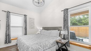 Ein Schlafzimmer mit Bett, Nachttisch, Lampe, Fenster mit Jalousien, gemusterten Vorhängen und einer Kristallleuchtre.