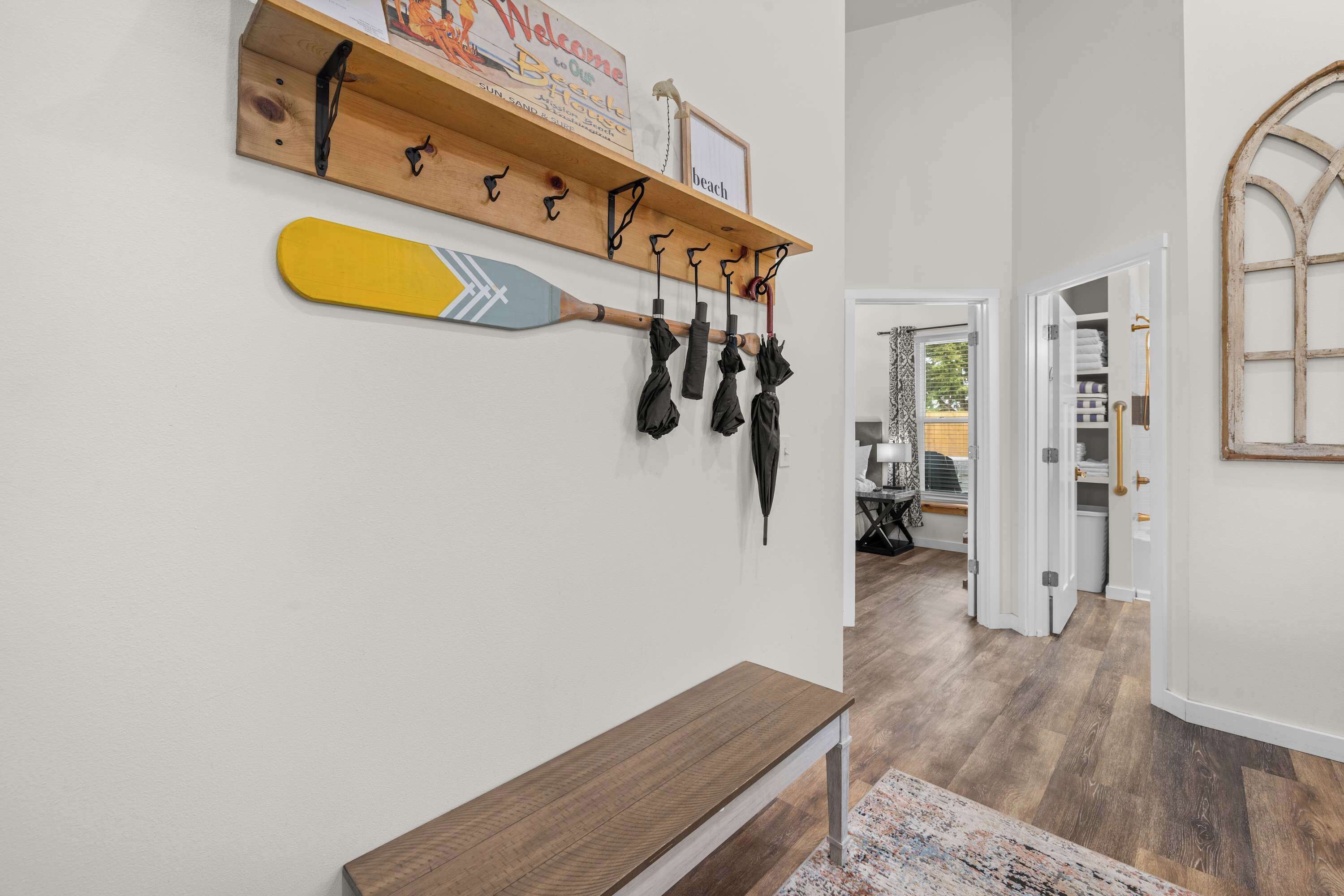 A hallway with a wooden bench, a wall-mounted rack with umbrellas, and a door leading to a room with a bed and a window.