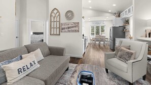 Ein modernes Wohnzimmer mit einem grauen Sofa, einem weißen Sessel und einem Couchtisch. An der Wand befinden sich eine Uhr und ein Schild mit der Aufschrift 'Bed & Breakfast'.