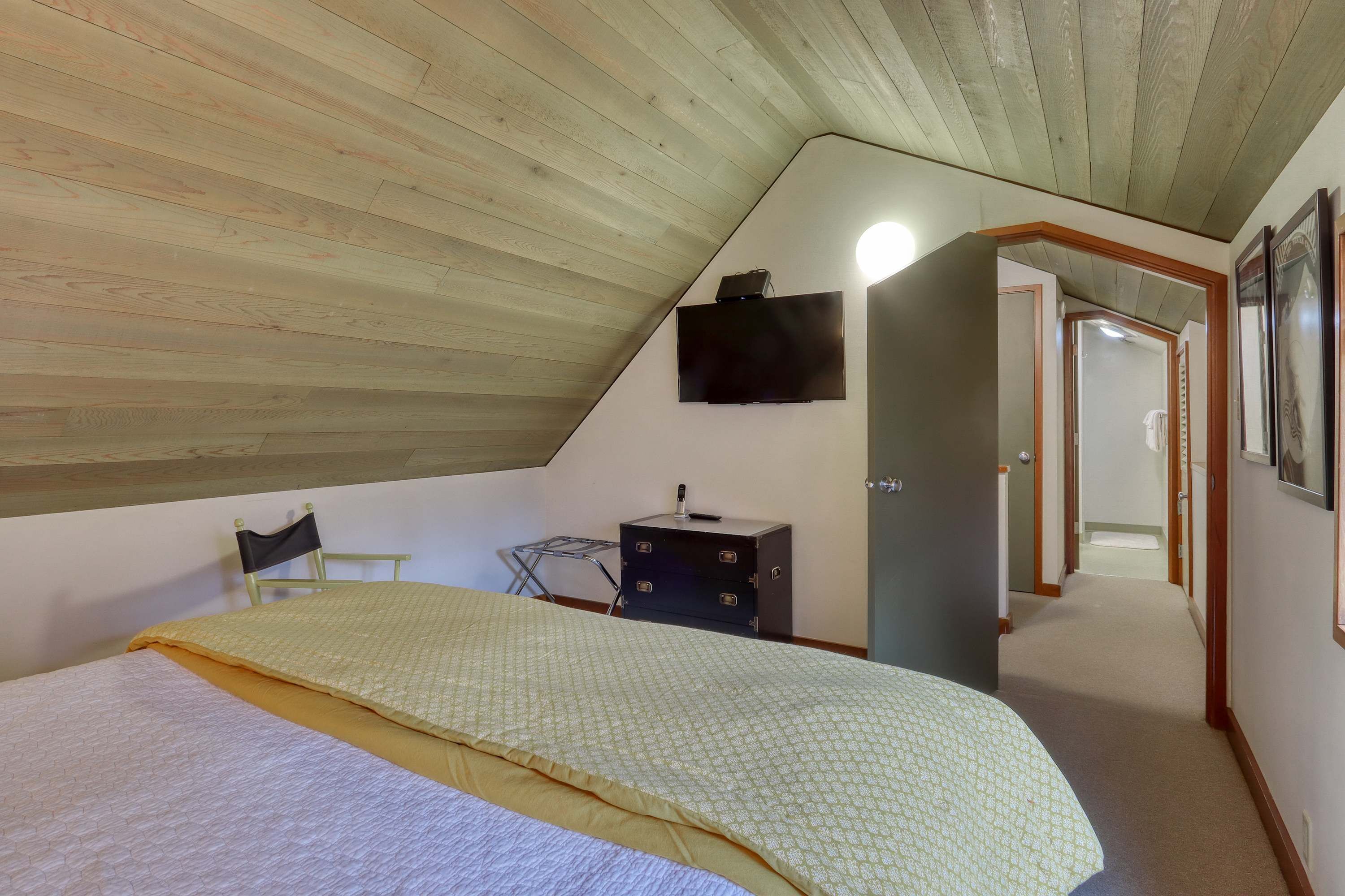 Ein Schlafzimmer mit schrägem Dach, einem Bett mit gemustertem Überwurf, einem Nachttisch mit Lampe, einem an der Wand befestigten Fernseher und einem Flur, der in ein weiteres Zimmer führt.